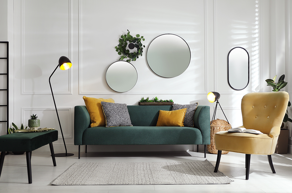 Moderni šviesių spalvų svetainė su žalia sofa, geltonomis pagalvėlėmis ir apvaliais veidrodžiais ant sienos.