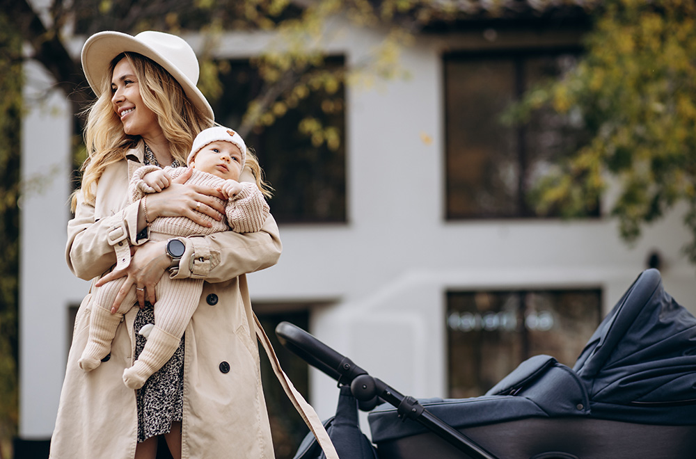 Moteris laiko kūdikį ant rankų, eidama šalia vežimėlio prie pastato.